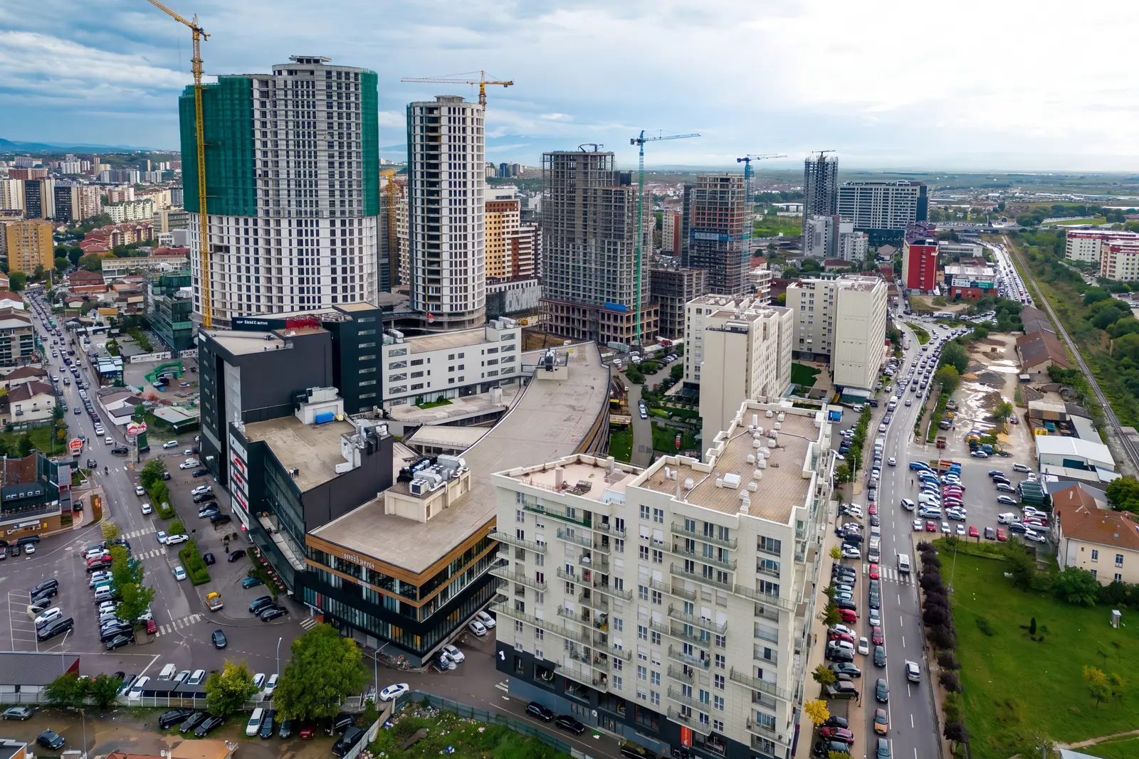 An aerial view of the city mid-construction: tower cranes over half-finished apartment towers, older streets below and open country on the horizon